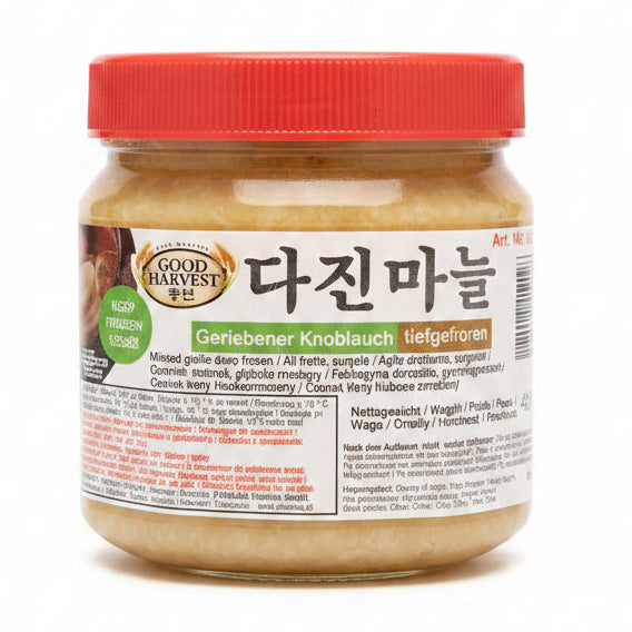 A hand holding a jar of Good Harvest Geriebeiner Knoblauch (garlic) with a red lid and a white label, set against a blurry indoor background.