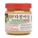 A hand holding a jar of Good Harvest Geriebeiner Knoblauch (garlic) with a red lid and a white label, set against a blurry indoor background.