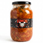 A large glass jar of Kimchi Korean fermented cabbage on a white refrigerator shelf. The jar has a black lid and a black label with red and white text reading 'KIMCHI' and a cartoon cow graphic.